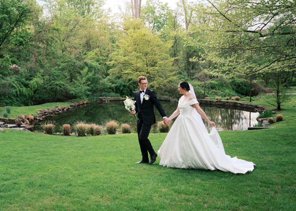 A bride and groom walkacross a green lawn during the golden hour, he in a tux, she in her off-the-shoulder wedding ball gown and cathedral veil