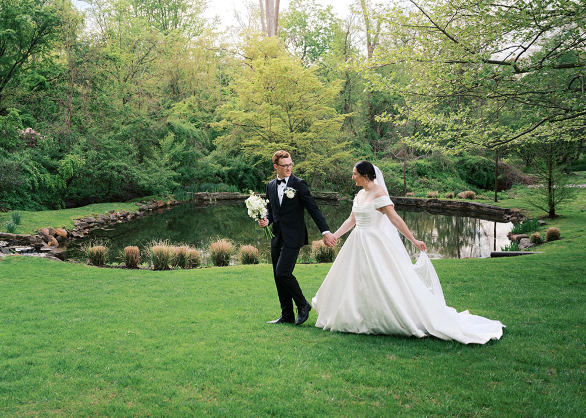 A bride and groom walkacross a green lawn during the golden hour, he in a tux, she in her off-the-shoulder wedding ball gown and cathedral veil
