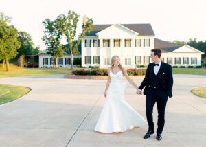 A bride and groom walk hand in hand smiling in the late afternoon sunlight. The bride wears a custom fit and flare wedding gown, the groom a tux