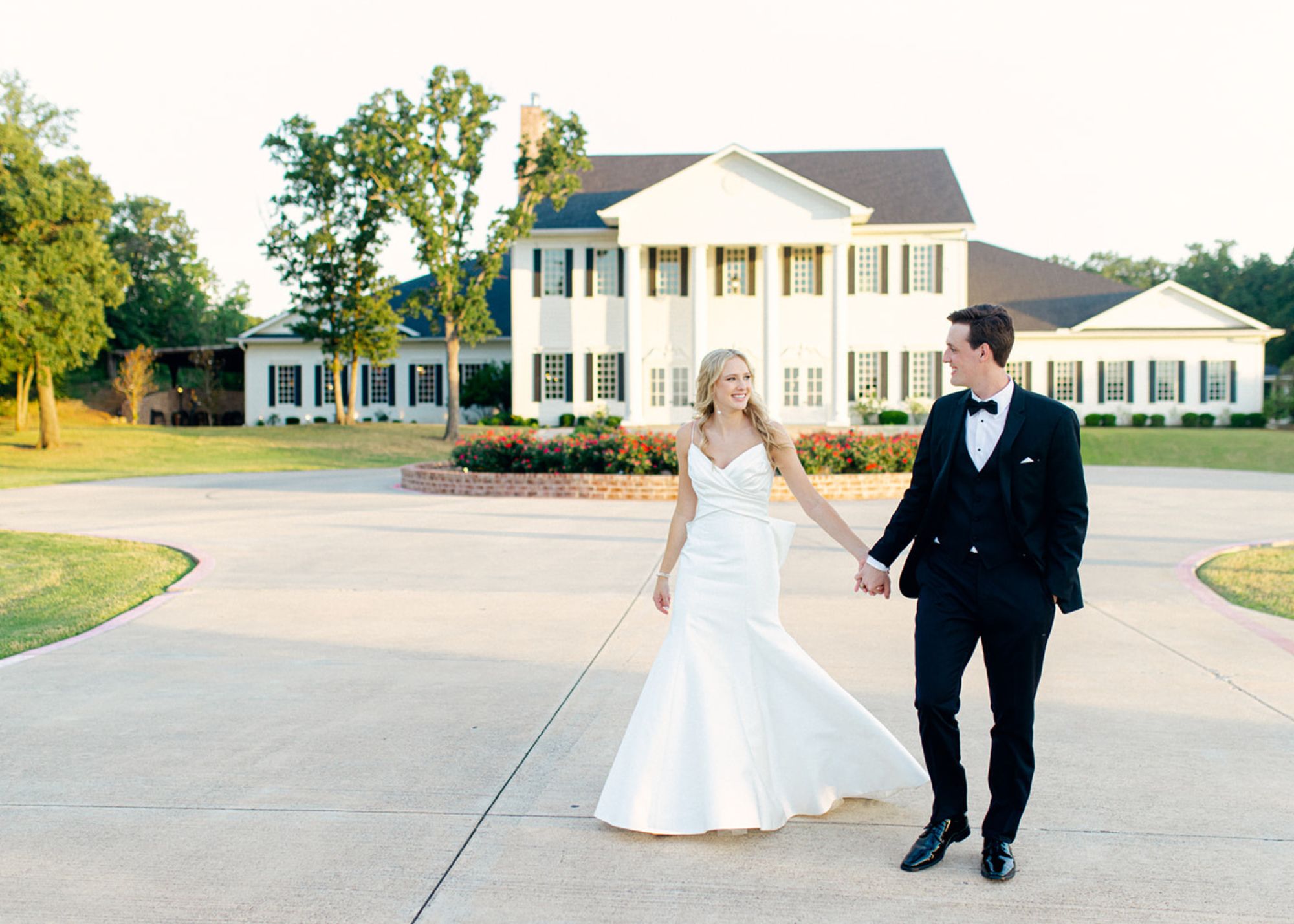 A bride and groom walk hand in hand smiling in the late afternoon sunlight. The bride wears a custom fit and flare wedding gown, the groom a tux