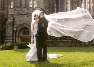 A bride and groom kiss outside before the church. the bride's veil flies in the sunshine, she wears a custom fit and flare wedding dress.