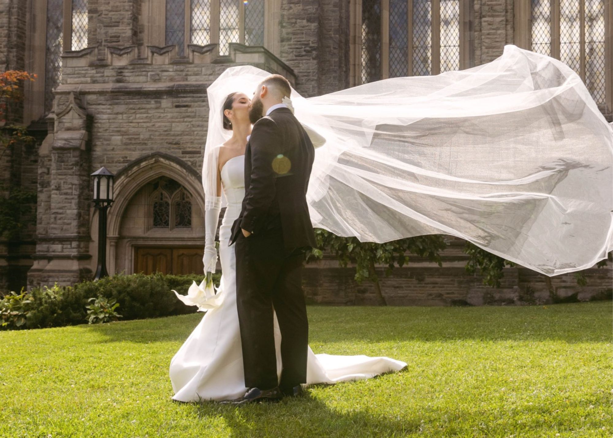 A bride and groom kiss outside before the church. the bride's veil flies in the sunshine, she wears a custom fit and flare wedding dress.