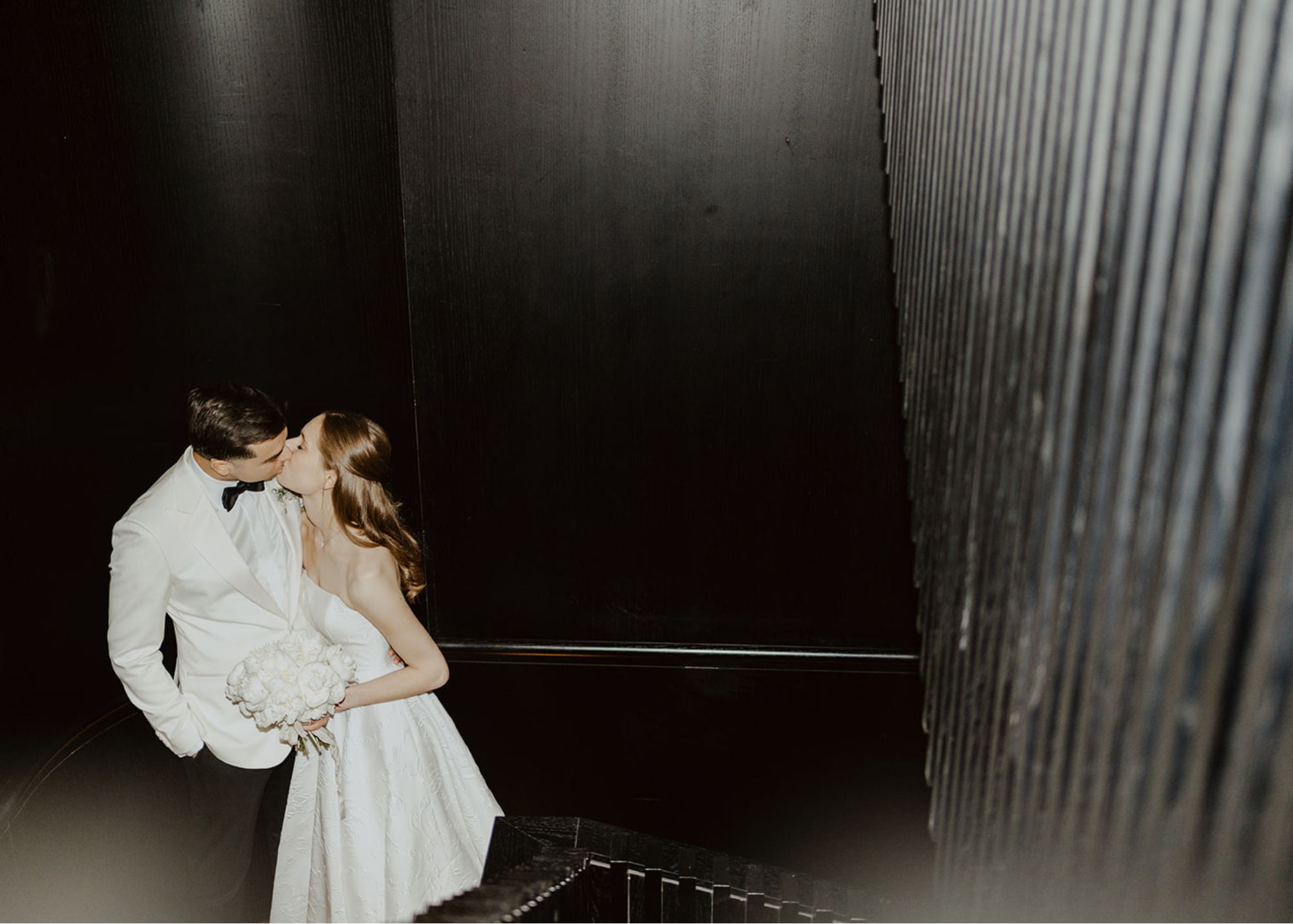 a bride and groom kiss in a dark wood stairwell. He wears a white diner jacket, she a floral Jacquard ball gown wedding dress