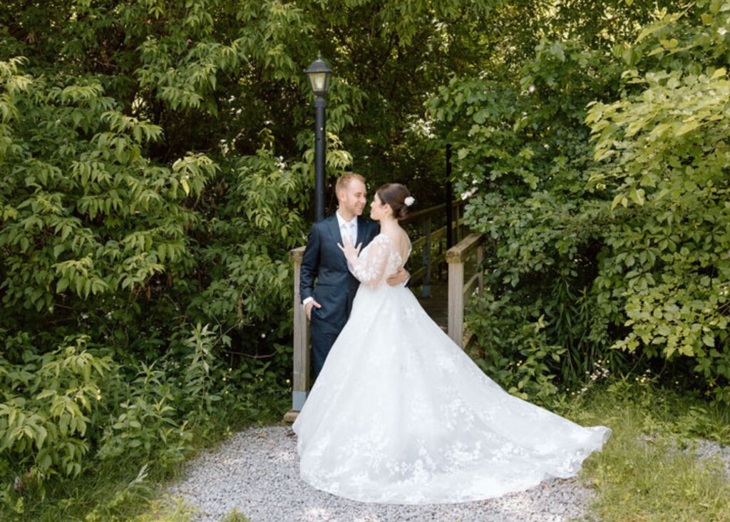 a bride and groom stand, holding one another an lush green wooded glen. The bride wears a floral ball gown wedding dress with long sleeves.