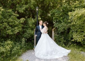 a bride and groom stand, holding one another an lush green wooded glen. The bride wears a floral ball gown wedding dress with long sleeves.