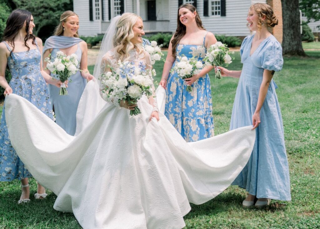 Bridesmaids in blue floral dresses holding bride's dramatic Lea-Ann Belter ballgown train in front of historic white colonial hous