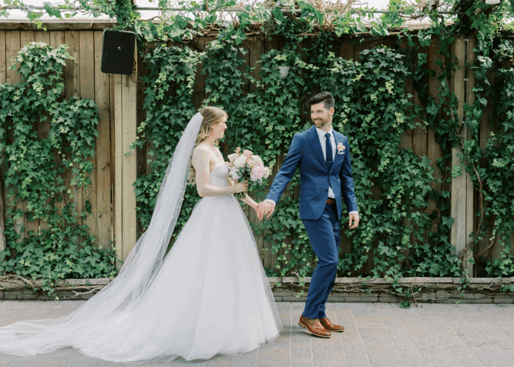 Bride in Lea-Ann Belter tulle ballgown with cathedral veil holding hands with groom in navy suit against ivy-covered wooden fence