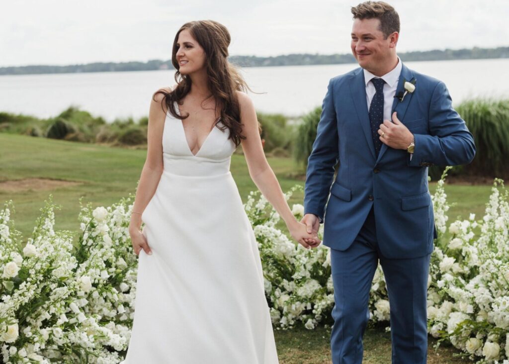 Newlyweds walking hand-in-hand past white floral arrangements at waterfront venue, bride in V-neck Lea-Ann Belter gown with train