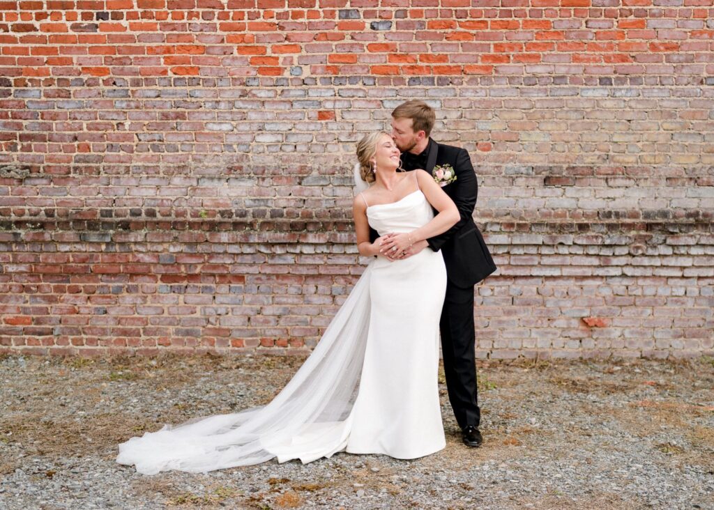 Groom embracing bride from behind against rustic brick wall, bride wearing sleek Lea-Ann Belter gown with draped neckline and dramatic train