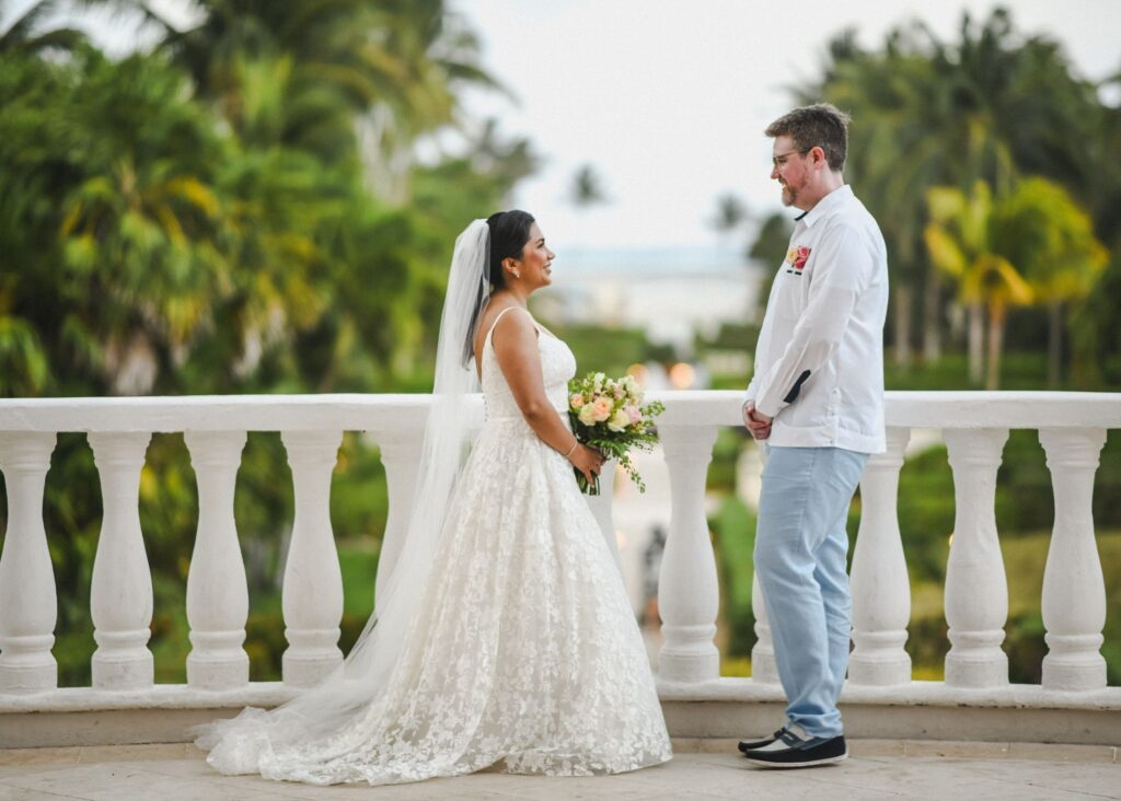 Bride and groom facing each other on white balustrade terrace with tropical palm trees, bride in lace Lea-Ann Belter A-line wedding dress with veil