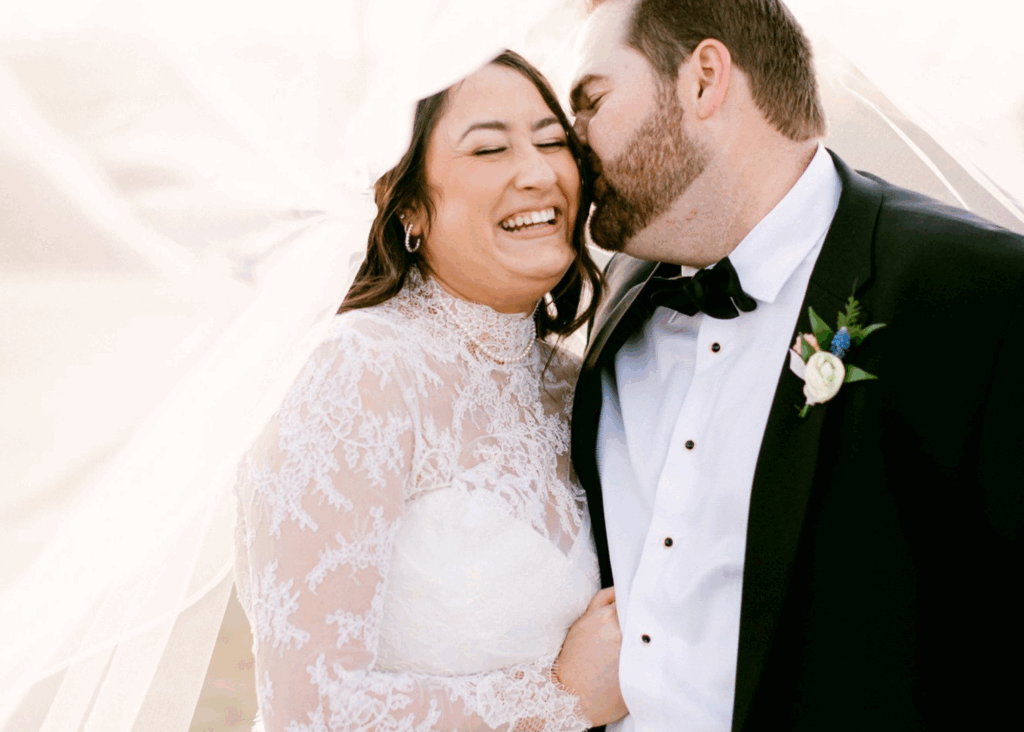 Groom kissing bride's cheek under flowing veil, bride laughing in Lea-Ann Belter gown with high-neck lace long sleeves
