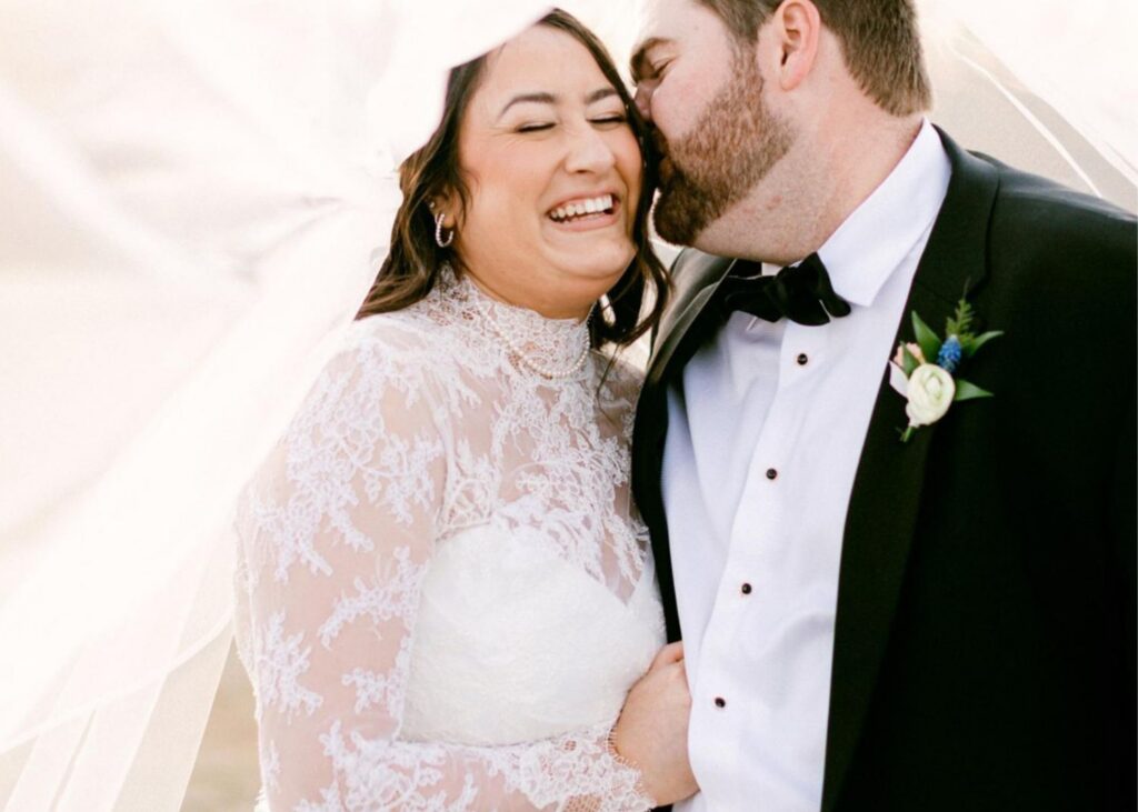 Groom kissing bride's cheek under flowing veil, bride laughing in Lea-Ann Belter gown with high-neck lace long sleeves