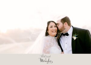 Carter wearing her Astrid & Mercedes Aura gown and Paradise lace topper, smiling as her groom Connor kisses her cheek during their March wedding in Winston-Salem