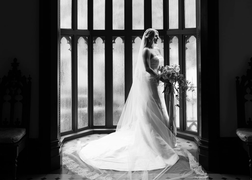 Black and white portrait of bride in strapless Lea-Ann Belter ballgown with cascading train standing by Gothic arched windows holding bouquet