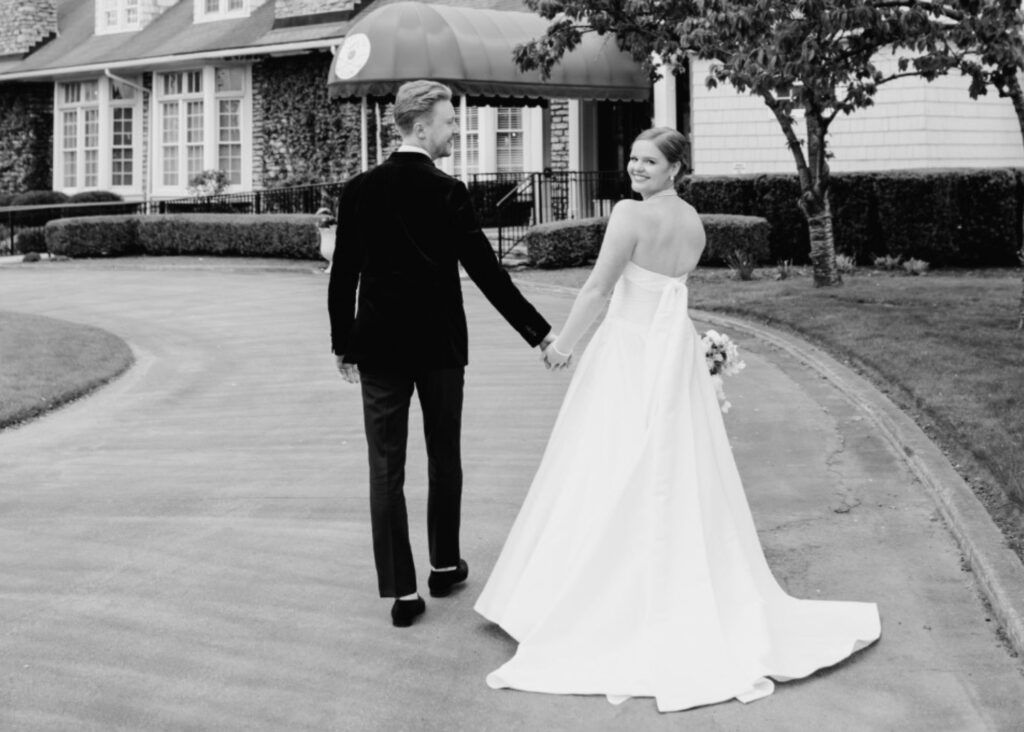 Black and white photo of newlyweds walking hand-in-hand at country club, bride in classic Lea-Ann Belter gown with train and tails