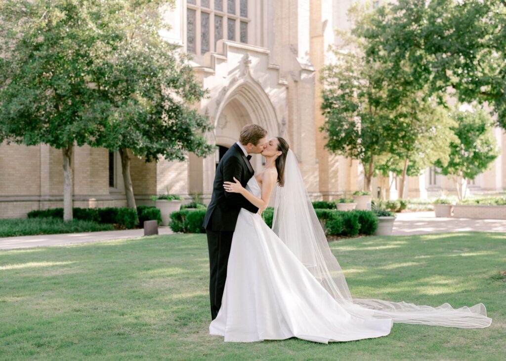 Bride and groom kissing on manicured lawn in front of Gothic church, bride wearing Lea-Ann Belter gown with cathedral veil