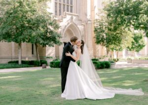 Bride and groom kissing on manicured lawn in front of Gothic church, bride wearing Lea-Ann Belter gown with cathedral veil