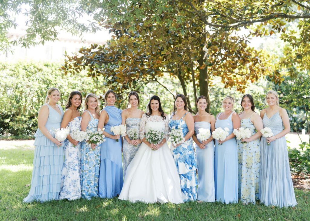 Bride in strapless Lea-Ann Belter ballgown with bridesmaids in blue and white dresses holding white bouquets under trees