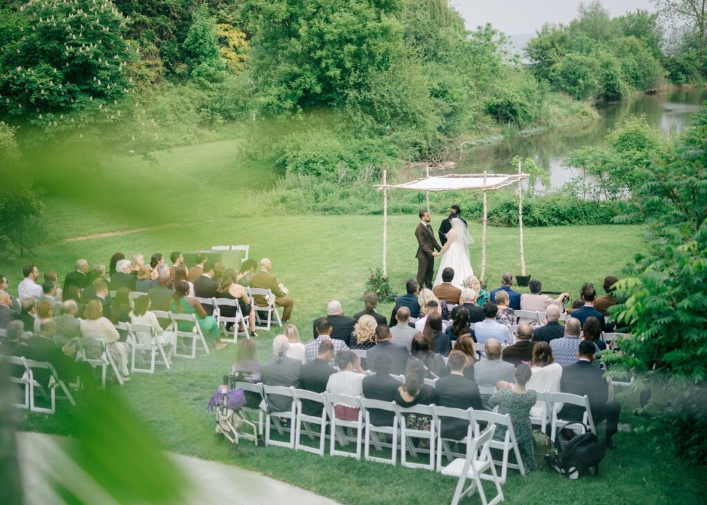Outdoor riverside wedding ceremony with guests seated on lawn, couple exchanging vows under a chuppa bride in Lea-Ann Belter gown