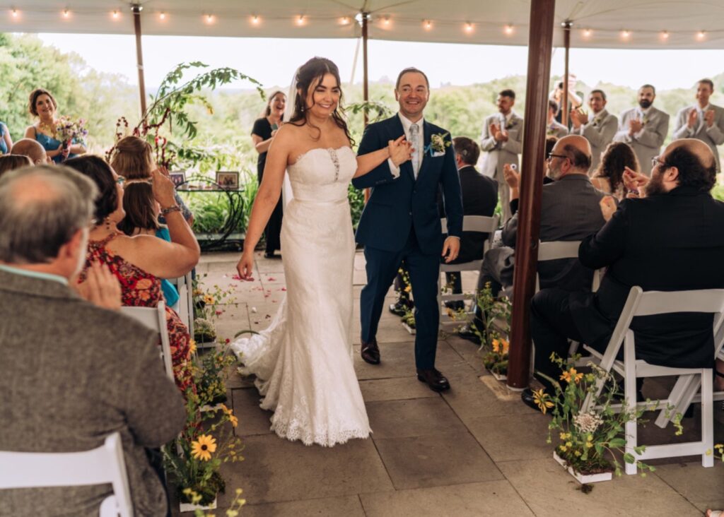 Newlyweds walking down ceremony aisle as guests applaud, bride in strapless lace gown with train at outdoor tented wedding