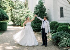 Groom twirling bride in strapless floral jacquard ball gown on gravel path in front of white painted brick building