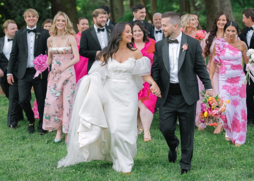 Newlyweds walking with wedding party through garden, bride in off-shoulder Lea-Ann Belter gown with dramatic shrug