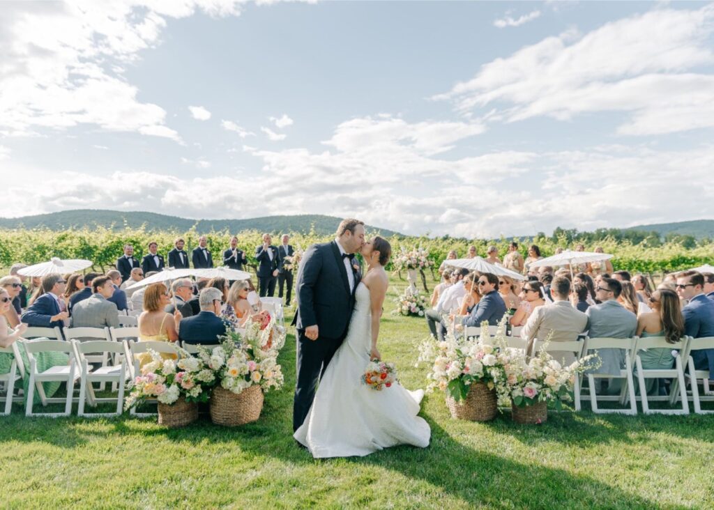 Bride and groom sharing first kiss at vineyard ceremony with mountain backdrop, bride in classic Lea-Ann Belter ballgown, guests with parasols