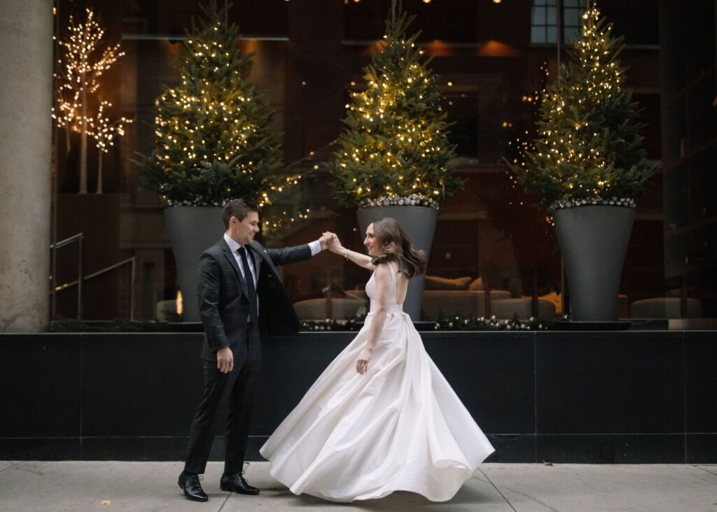 Bride twirling in elegant Lea-Ann Belter ballgown with flowing skirt as groom extends hand, illuminated trees in background