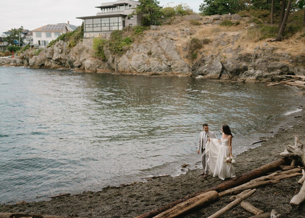 Newlyweds walking along rocky Pacific Northwest beach with driftwood, bride wearing flowing Lea-Ann Belter gown