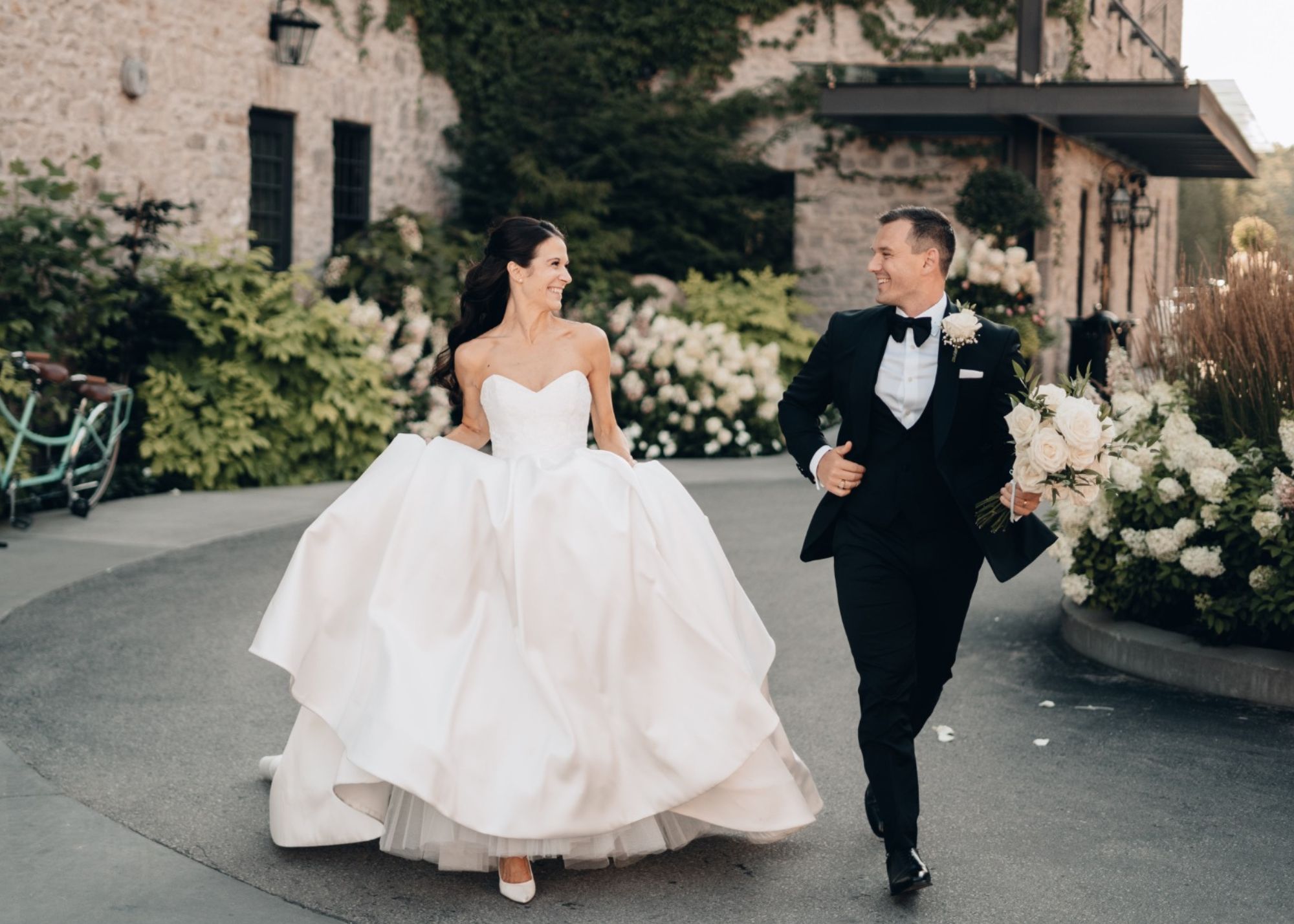 A bride and groom run joyfully in front of there mansion venue. The bride wears a custom ball gown wedding dress from Lea-Ann Belter Toronto