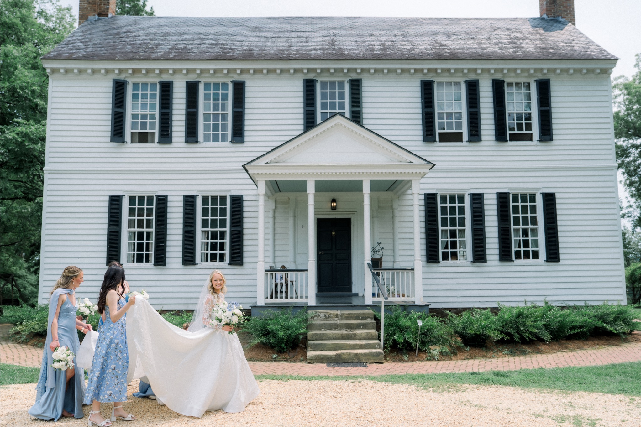 Bridesmaids helping bride in a elegant ball gown wedding dress with long lace sleeves in front of classic white colonial house