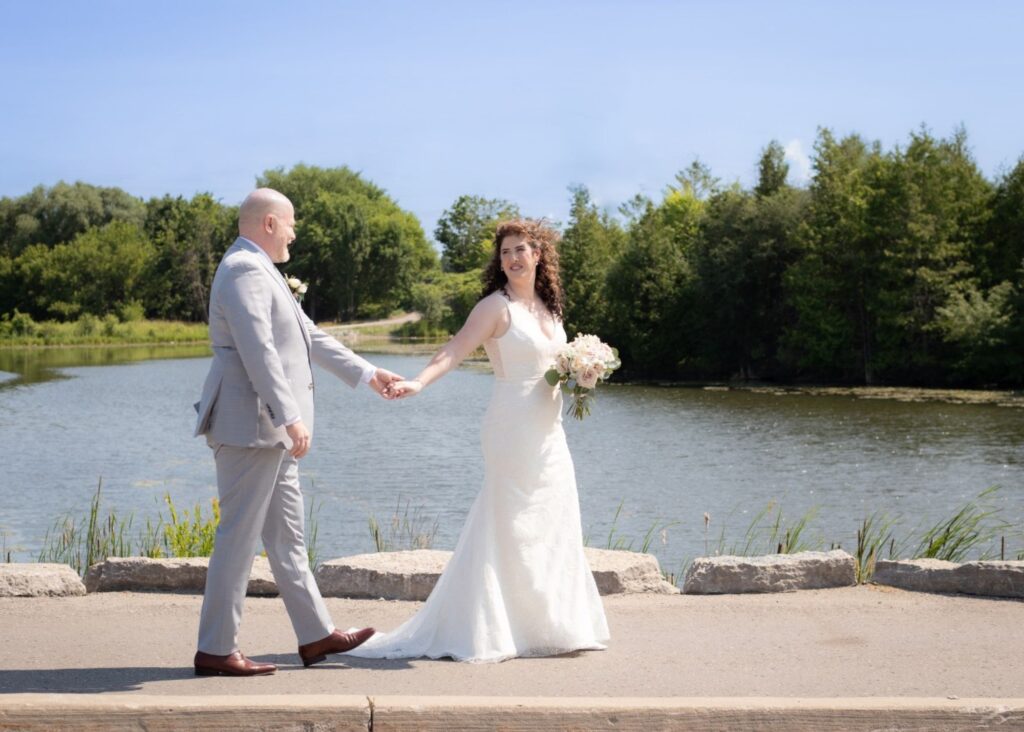 A bride and groom walk hand in hand by water in the sun The bride wears her beaded, sleek wedding dress by Lea-Ann Belter Bridal