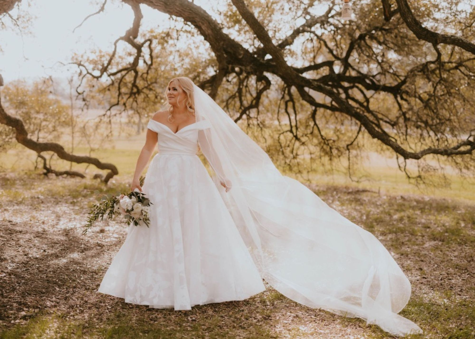 Bride Delaney in a classic off-the-shoulder white ball gown wedding dress with cathedral veil, holding a bouquet under sprawling oak trees in Austin, Texas