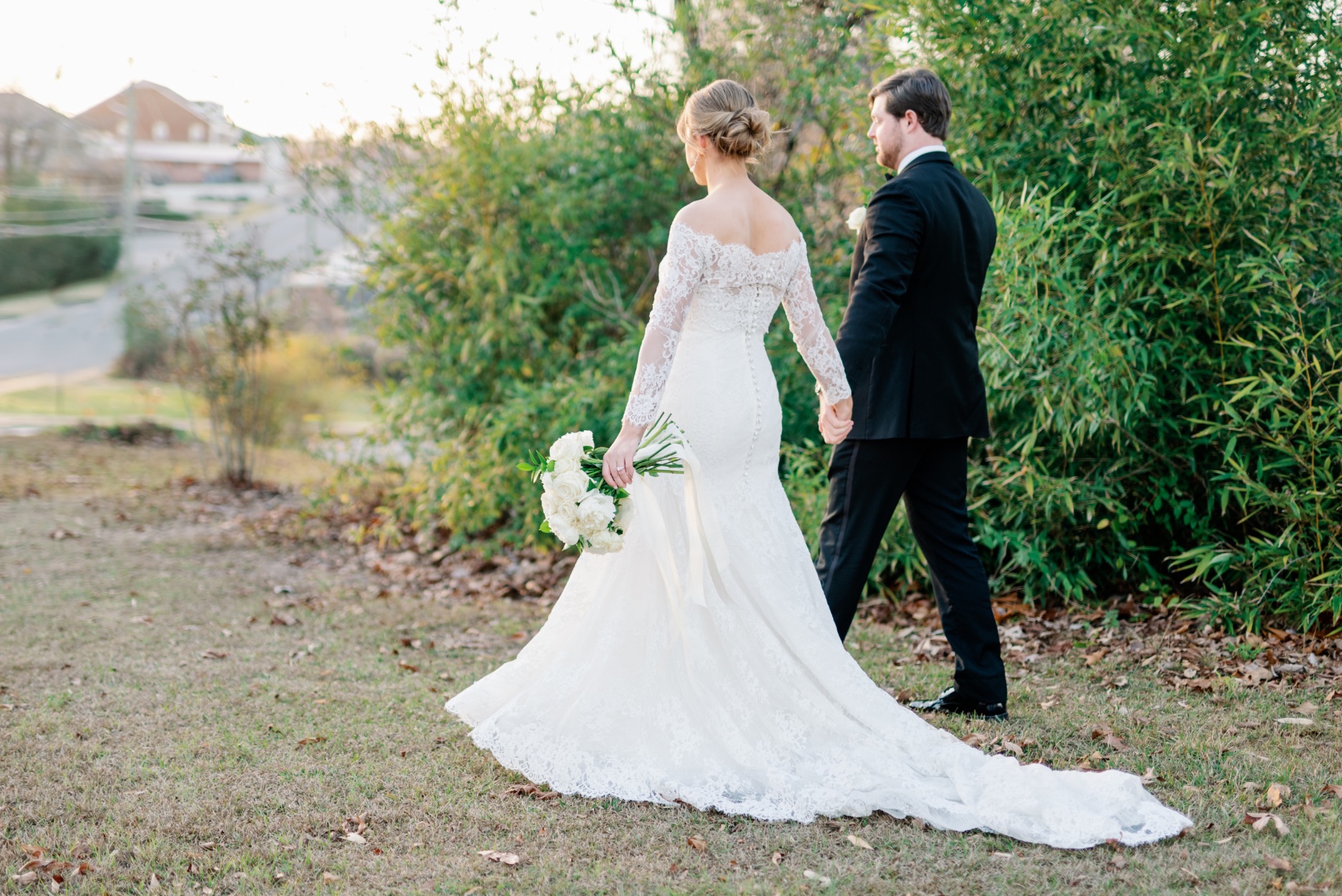 Bride wearing Lea-Ann Belter long-sleeved lace gown with flowing train holding groom's hand in outdoor garden setting