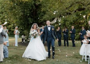 A delighted bride and groom walk down the aisle amidst tossed petals. The bride wears a ball gown wedding gown with tulle sleeves.