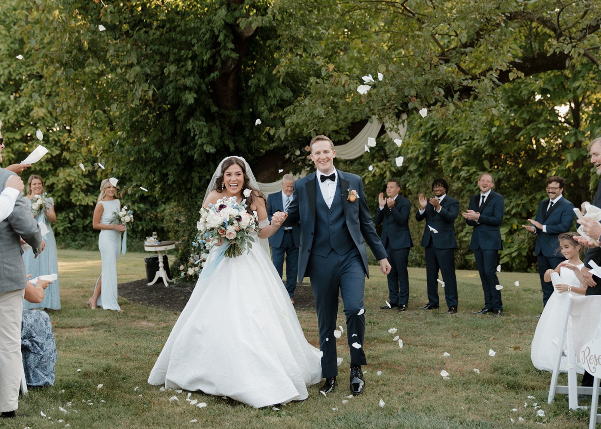 A delighted bride and groom walk down the aisle amidst tossed petals. The bride wears a ball gown wedding gown with tulle sleeves.