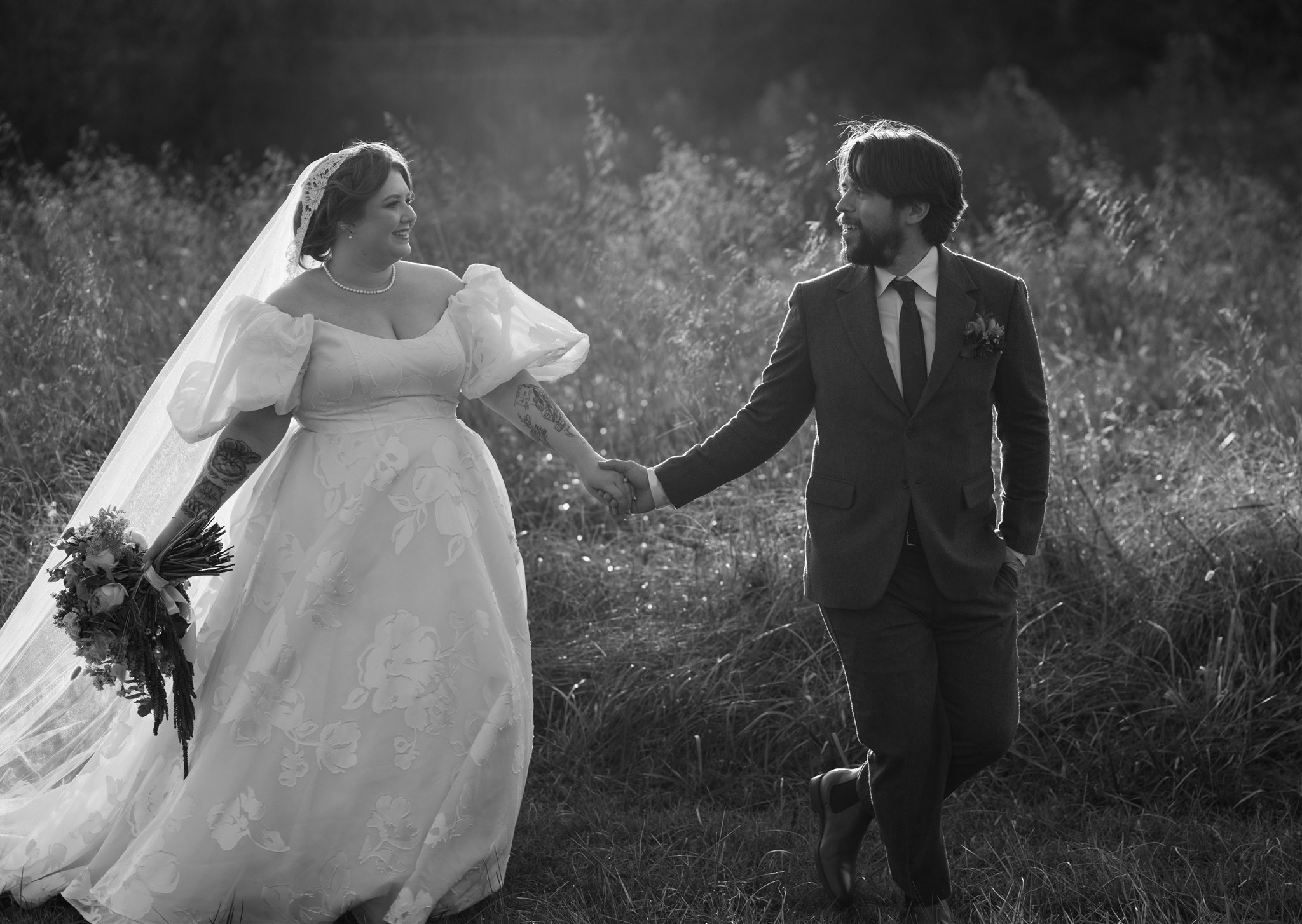 Black and white wedding photo of bride wearing Lea-Ann Belter Esperanza gown featuring romantic puff sleeves and full ballgown skirt, walking hand-in-hand with groom through wildflower field