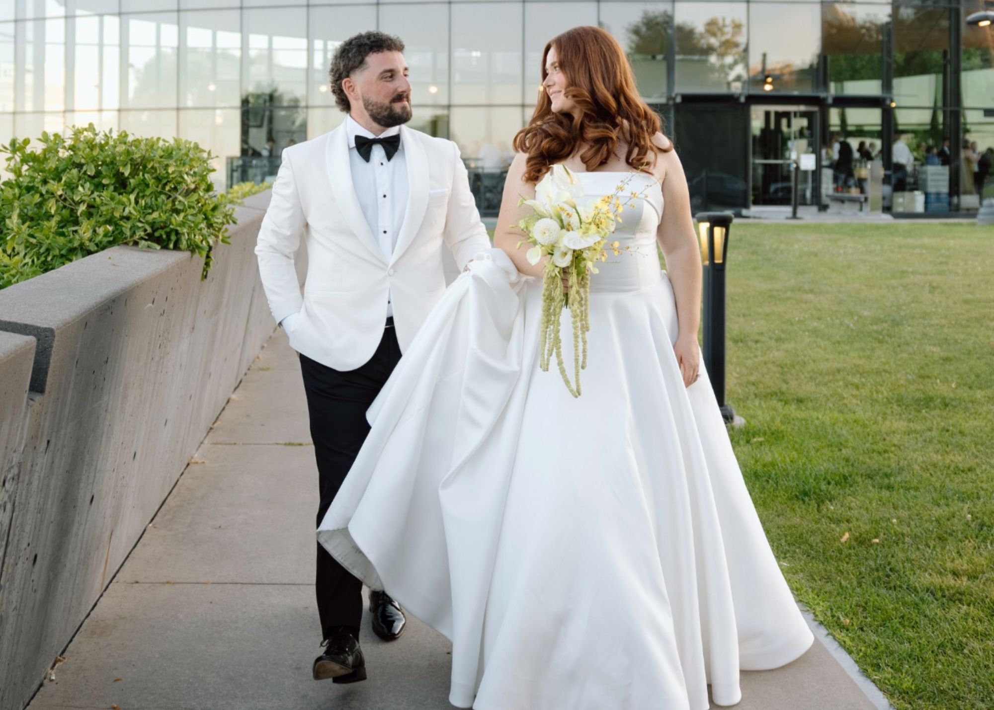 Jaqueline and Matthew walk outside together, Matthew holds the train of his bride's ball gown wedding dress