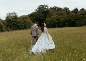 Bride wearing custom pale blue ballgown by Lea-Ann Belter holds calla lily bouquet while walking through wildflower field in Ireland with groom