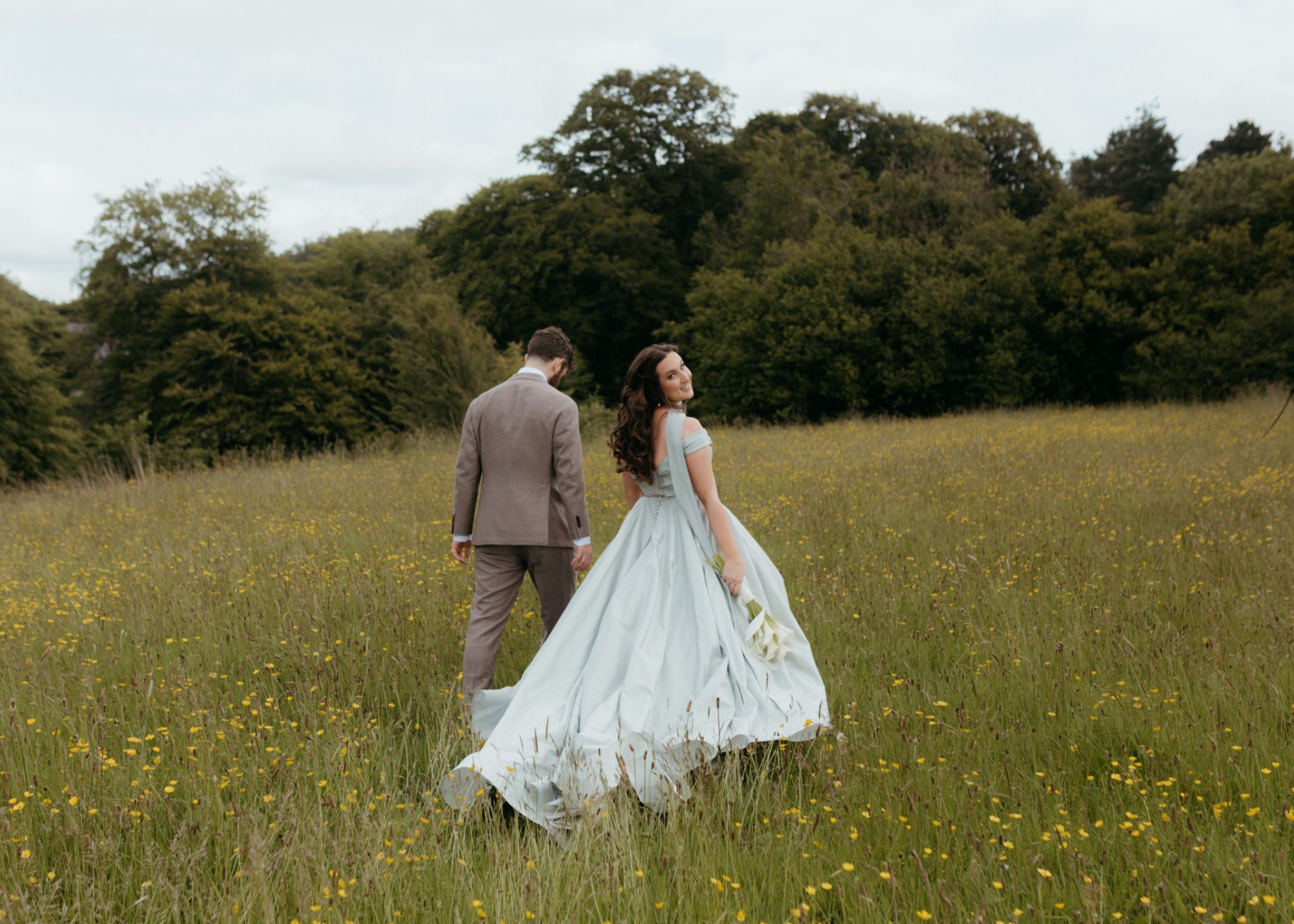 Bride wearing custom pale blue ballgown by Lea-Ann Belter holds calla lily bouquet while walking through wildflower field in Ireland with groom