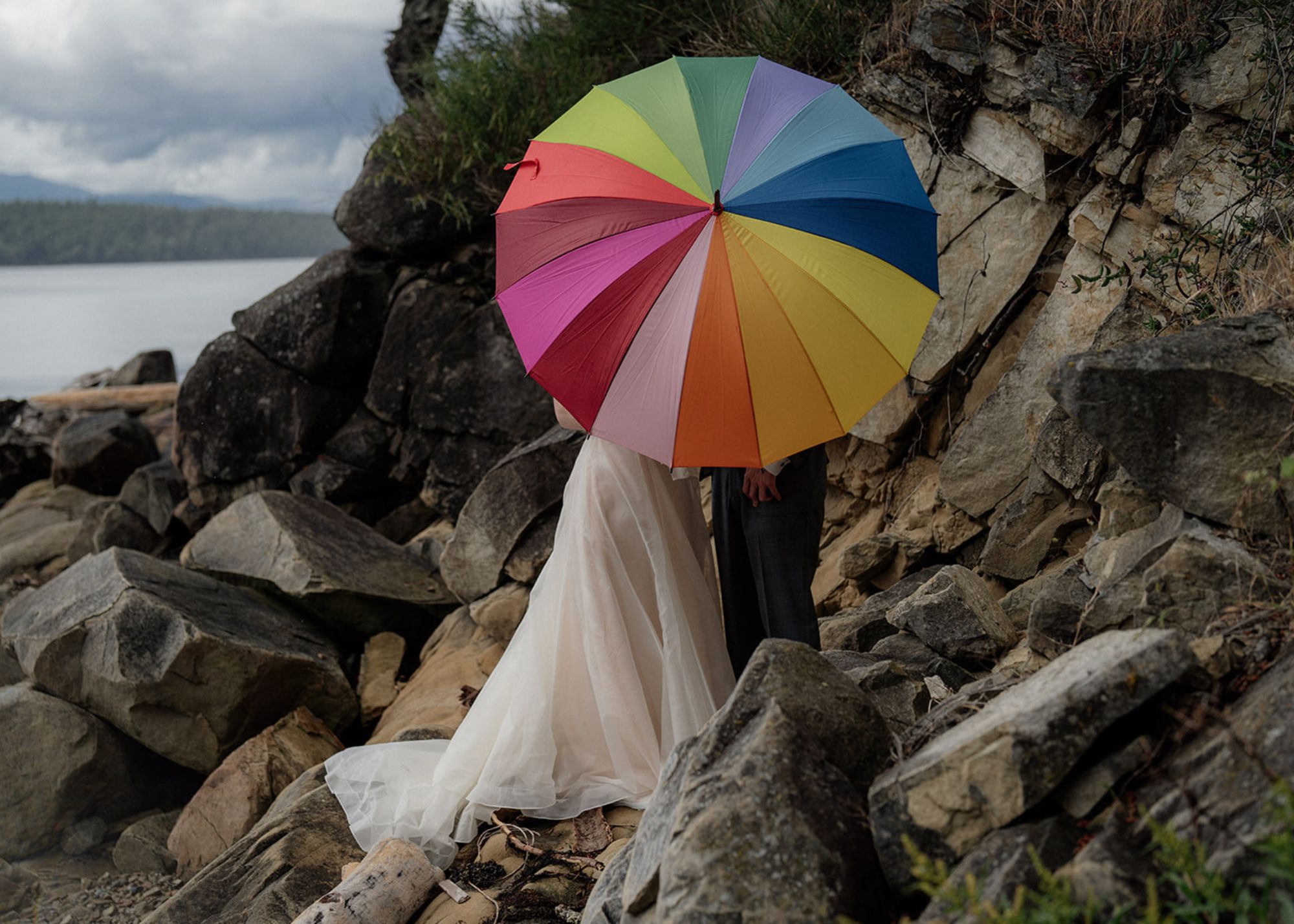 a bride and groom kiss while standing on a rock by the bay, their faces covered by a rainbow umbrella. The bride wears bridal separates by Lea-Ann Belter Bridal