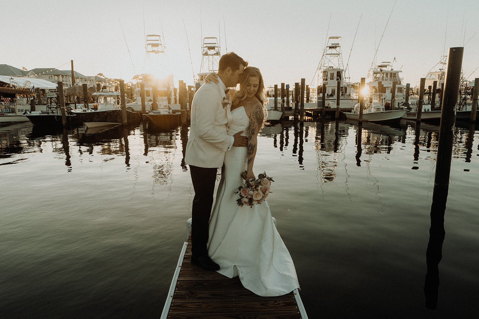 Bride in fit and flare Lea-Ann Belter wedding dress with dramatic train embracing groom at marina dock during golden hour sunset