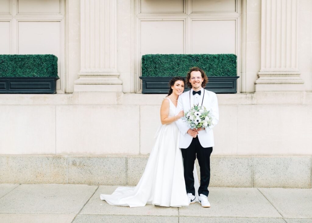 Bride in classic A-line wedding dress with sleeveless bodice holding anemone bouquet with groom in white tuxedo jacket