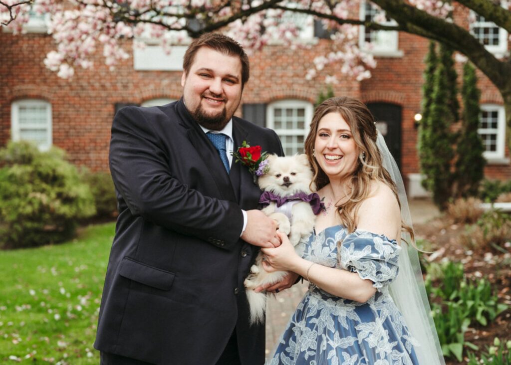 Bride Olivia in custom blue Lea-Ann Belter lace wedding gown with groom Krystian holding their Pomeranian Nimbus in front of brick building