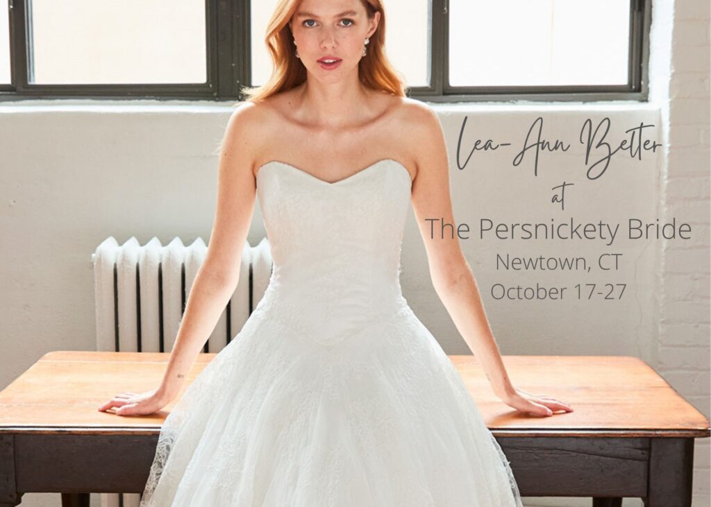 Bride wearing a strapless ivory ballgown wedding dress with textured lace and tulle skirt, seated on a wooden table in a bright studio setting. Text reads 'Lea-Ann Belter at The Persnickety Bride, Newtown, CT, October 17-27'.