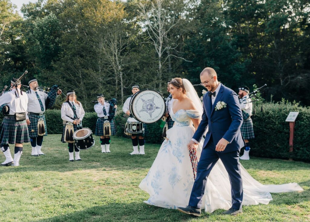 Bride Ella in a custom blue and white bridal ball gown with off-shoulder details walks hand-in-hand with groom Jay in a navy suit, both smiling as they pass a traditional Scottish pipe and drum band performing on the lawn at their Massachusetts wedding