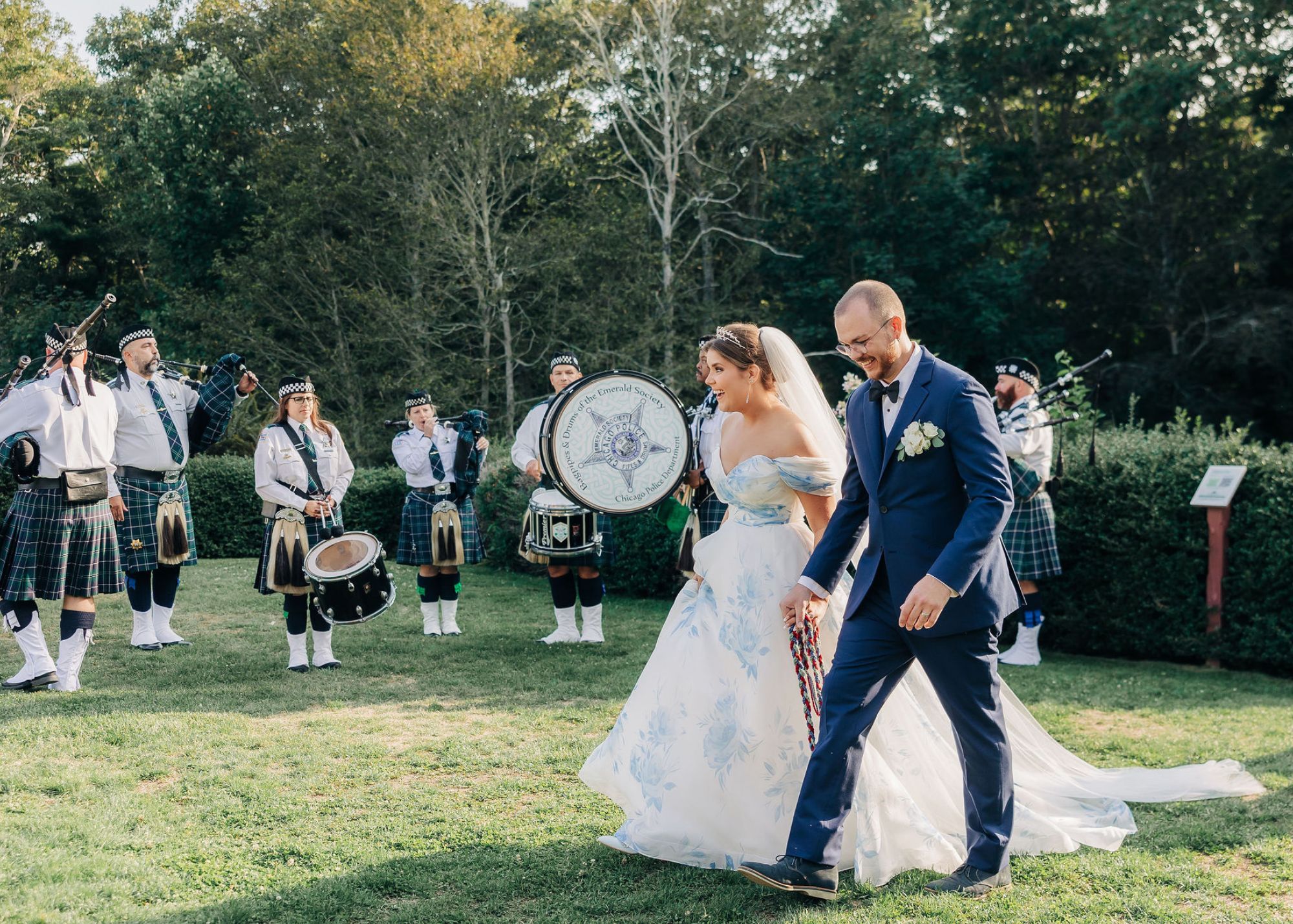 Bride Ella in a custom blue and white bridal ball gown with off-shoulder details walks hand-in-hand with groom Jay in a navy suit, both smiling as they pass a traditional Scottish pipe and drum band performing on the lawn at their Massachusetts wedding