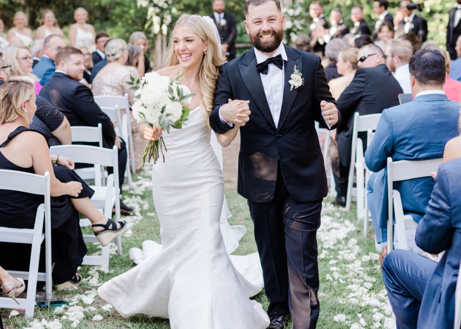 A just married bride and groom walk happily down the aisle, arm in arm. The bride is wearing her custom lace fit and flare wedding dress
