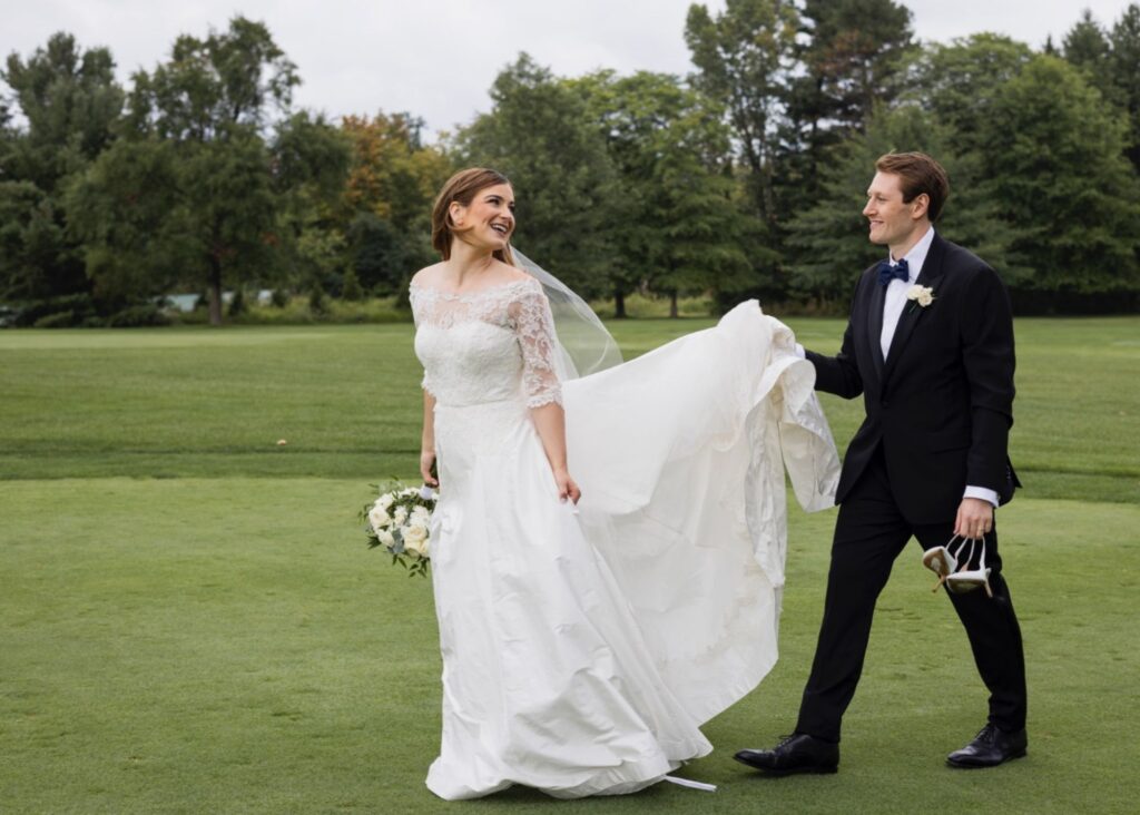 Real bride Katie walks with her husband Karl across a green lawn. He holds her a-line wedding dress's train up so it won't get wet.