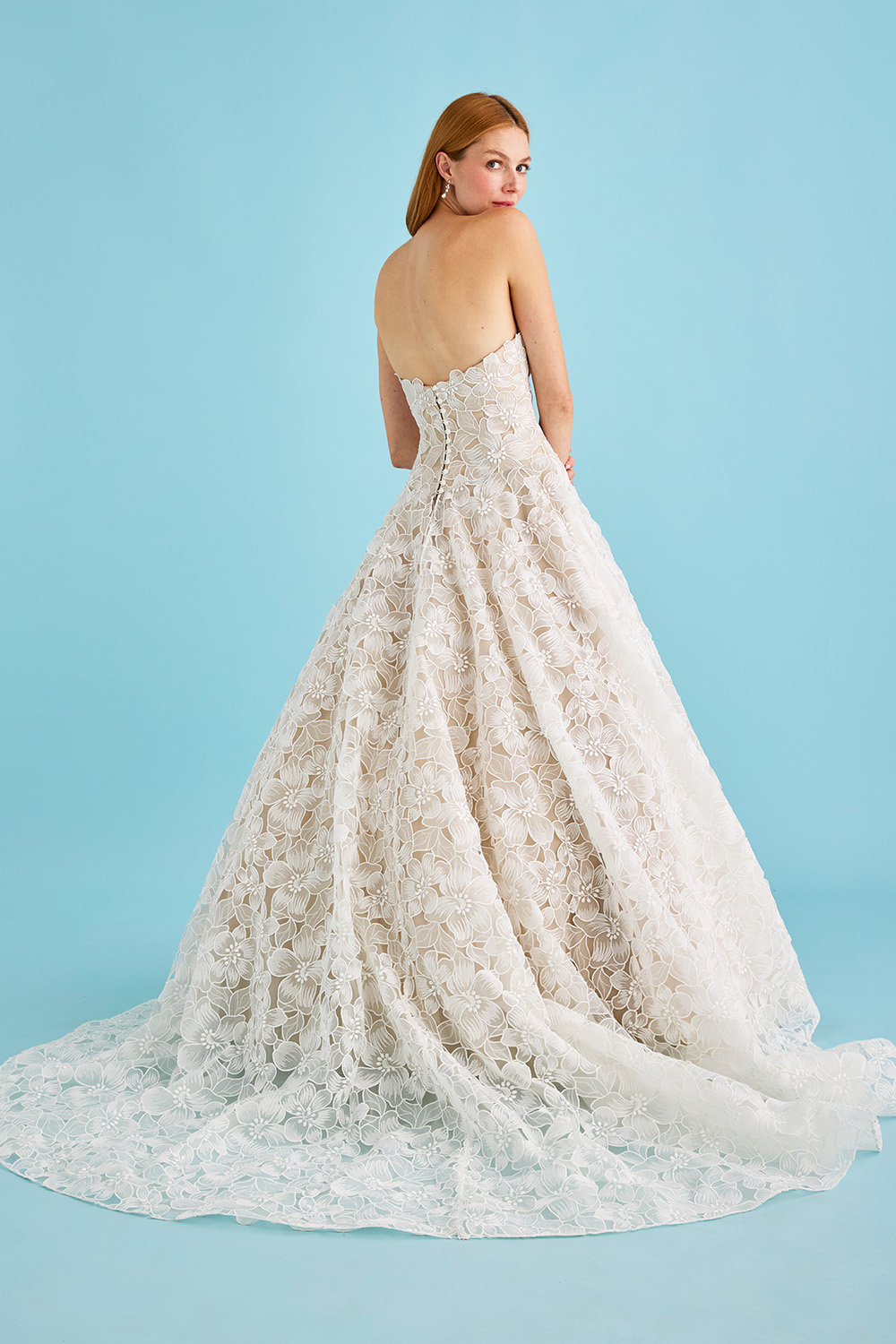 A model looks over her shoulder wearing the Luanne strapless ball gown wedding dress of cut organza anemone flowers, it's dramatic, sheer train veils the floor.
